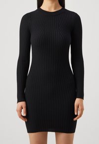 Kvinna iklädd en åtsittande svart ribbad stickad klänning med långa ärmar och rundad hals, visad från halsen till mitten av låret mot en enfärgad bakgrund.