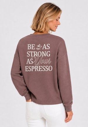 Kobieta o krótkich blond włosach, nosząca brązową bluzę z napisem "Be as strong as your espresso" na plecach, w połączeniu z białymi spodniami.