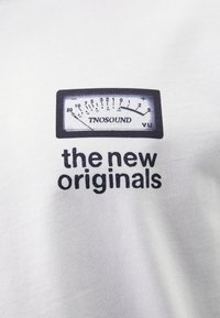 Hvit bomull T-skjorte med grafikk av en vintage VU-måler med teksten "TNOSOUND" over og "de nye originalene" under i svart skrift.