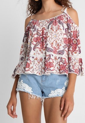 Blusa bianca off-shoulder con stampa floreale e paisley, scollo elastico e orlo arricciato. Abbinata a pantaloni bermuda in denim chiaro con dettagli in pizzo.