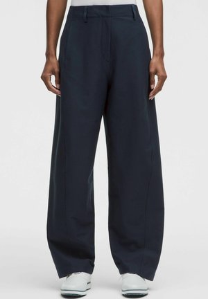 Persona che indossa pantaloni neri larghi e sneaker bianche, in piedi davanti a uno sfondo semplice.