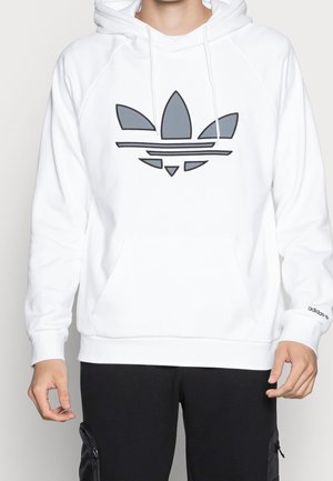 Persoon draagt een witte hoodie met groot grijs Adidas trefoil-logo op de borst en een zwarte broek met ritszakken.