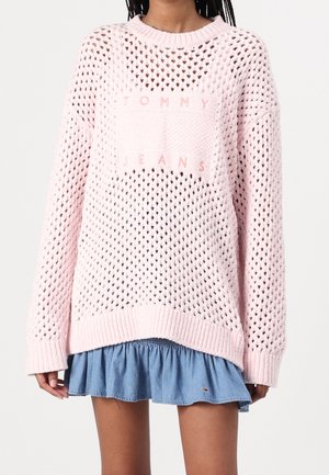 Person iført en lyserød åbent strikket sweater med "Tommy Jeans"-logo over en kort blå flæsekjol.