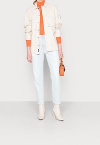 Camisa chaqueta oversized beige, cuello alto naranja, jeans de cintura alta de color azul claro y botines crema. Un pequeño bolso naranja completa el conjunto.