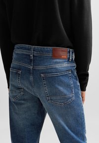 Mörkblå jeans i denim med mjuk yta, utrustade med bakfickor och en brun läderlapp märkt "BOSS" vid midjan.