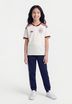 Smilende barn stående iført hvid spansk fodboldtrøje med røde accenter, mørkeblå træningsbukser med hvide striber og hvide sneakers.