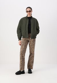 Casaco bomber verde-oliva, blusa preta, calças com padrão animal em tons de castanho e sapatos pretos. Ajuste slim, tecido texturizado e fechos em zíper no casaco.