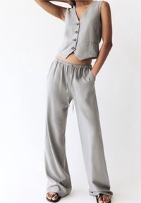 Gilet en lin gris avec un col en V et cinq boutons, associé à un pantalon gris ample à taille haute avec une ceinture élastique et des poches latérales.