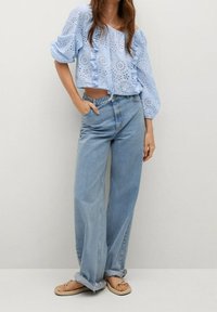 Vaaleansininen silmäreikäpusero, jossa on joustavat hihansuut ja röyhelöity etuosa, yhdistetty korkeavyötäröisiin vaaleansinisiin leveälahkeisiin farkkuihin ja beigen värisiin varvassandaaleihin.