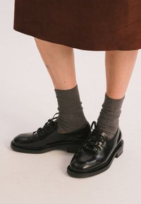 Scarpe in vernice nera con punte tonde e design con lacci, abbinate a calze a coste grigio scuro. La scarpa presenta un tacco basso e spesso.