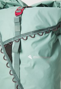Sac à dos vert en tissu durable avec des accents rouges contrastants, des bretelles réglables et un détail de couture circulaire. Logo de la marque affiché.