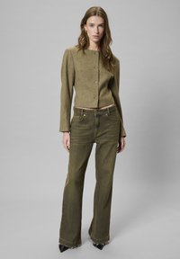 Oliven grøn, cropped, struktureret jakke med sølvknapper, parret med højtaljede, udvidede, slidte grønne jeans og sorte spidse hæle.
