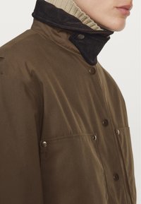 Braune äußere Jacke mit einem schwarzen Cordkragen, ausgestattet mit Druckknöpfen und zwei Vordertaschen. Der Stoff wirkt strukturiert und langlebig.