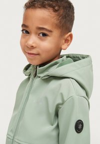 Lys grøn zip-up jakke med hætte, glat stof, med en cirkulær sort logo-patch på venstre ærme og et minimalistisk design.