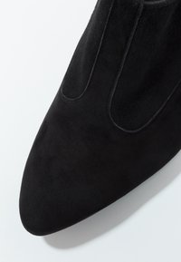 Botte de cheville en suède noir avec un bout pointu, dotée de panneaux lat éraux élastiques et d'une texture lisse et mate. Vue en gros plan de la partie supérieure.