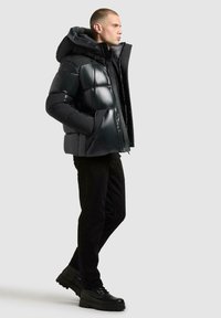 Zwarte pufferjas met een glanzende afwerking, oversized capuchon, rits aan de voorkant en zijzakken; gedragen over zwarte broek en chunky zwarte laarzen.