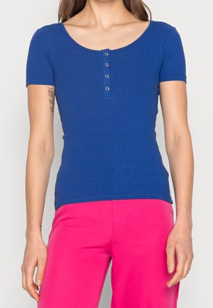 Sininen ribbineulottu lyhythihainen paita, jossa on pyöreä pääntie ja viisi painonappia keskellä. Yhdistetty kirkkaan pinkkien housujen kanssa.