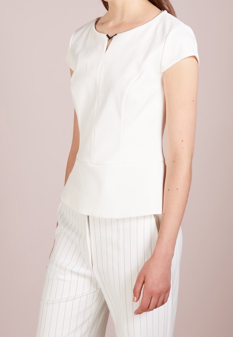 Haut blanc à manches courtes avec un col rond et une découpe en forme de V à l'avant, associé à un pantalon blanc à rayures. Tissu lisse et structuré.