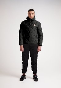 Svart pufferjacka med diamantquiltat mönster, hög krage och logo på bröstet, i kombination med svarta cargobyxor och svarta sneakers.