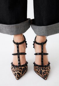 Tacones de aguja con estampado de leopardo y tiras negras con tachuelas. El diseño de punta afilada presenta un empeine texturizado y detalles de color contrastantes.