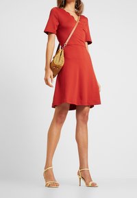 Žena v červených šatech s krátkým rukávem a vlnkovým výstřihem, žlutých podpatcích, velkých náušnicích ve tvaru kruhu a tkané crossbody tašce.