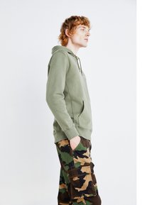 Felpa verde oliva con tasca frontale e coulisse regolabile, abbinata a pantaloni cargo mimetici con più tasche in verde e marrone.