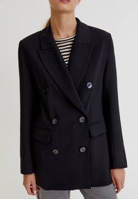 Femme portant un blazer noir croisé à quatre boutons, sur un haut rayé noir et blanc et un pantalon gris, sur un fond uni.