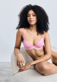 Różowy miękki biustonosz z koronkowymi wykończeniami wzdłuż dolnej krawędzi. Proste ramiączka i gładki materiał; modelka siedzi na teksturowanym, zaokrąglonym krześle.