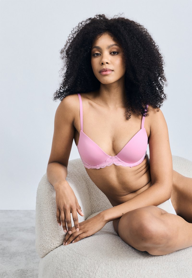 Różowy miękki biustonosz z koronkowymi wykończeniami wzdłuż dolnej krawędzi. Proste ramiączka i gładki materiał; modelka siedzi na teksturowanym, zaokrąglonym krześle.