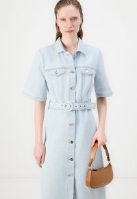 Lys blå denim skjortekjole med korte ermer, knapper nedover fronten og belte i midjen. Brun håndveske holdt i høyre hånd.