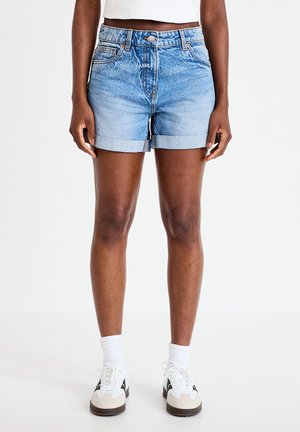 Osoba w jasnoniebieskich, podwiniętych szortach dżinsowych, białych trampkach, białych skarpetach do kostek oraz białym, krótkim topie, stojąca na białej podłodze.