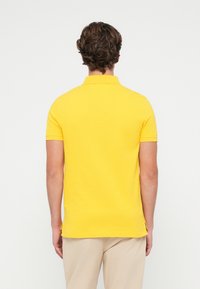 Gul kortermet polo skjorte med ribbestrikket krage, sidesplitt ved hemmen, og en glatt, myk tekstur. Bakre utsikt viser en tettsittende design.
