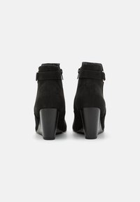 Bottines en cuir noir suédé avec un talon haut, dotées d'une fermeture éclair et de détails de sangle à la cheville. Texture lisse et design à bout rond.