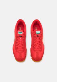 Red athletic shoes with a suede and textile upper, flat laces, gum rubber outsole, and white side accents. Green and white brand logo on tongue.