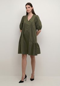 Oliņu zaļš puff-sleeve kleita ar V veida kaklu un daudzslāņu apakšmalu. Izgatavota no viegla auduma, ar maigu tekstūru un brīvi plūstošu siluetu.