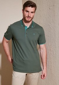 Zelena polo majica s kratkimi rokavi z turkiznimi detajli, zatesnjenim krojem, tremi gumbi in subtilnim logotipom na prsih. Gladka tekstura, prilegajoč dizajn.