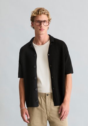 Junger Mann mit blondem Haar und Brille, der ein schwarzes kurzärmeliges Hemd über einem cremefarbenen Top und beigefarbenen Hosen vor einem einfarbigen Hintergrund trägt.