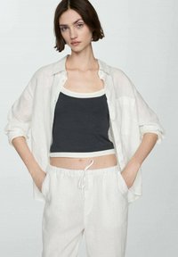 Mlada ženska, oblečena v belo odprto srajco čez črn crop top in bele hlače z vrvico, stoji s rokami v žepih na enostavni podlagi.
