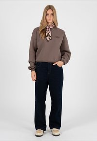 Frau mit langen blonden Haaren trägt ein braunes Sweatshirt, einen floralen Halsschal, dunkle Jeans mit weitem Bein und beige Turnschuhe, steht mit einer Hand in der Tasche.