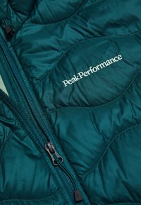 Giacca trapuntata turchese con zip nera e logo "Peak Performance" bianco ricamato sul petto.