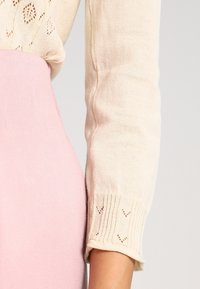 Jasno beżowy sweter z fakturowanym wzorem dzianiny, z ribbami na rękawach, obok częściowo widoczna jest miękka różowa spódnica.