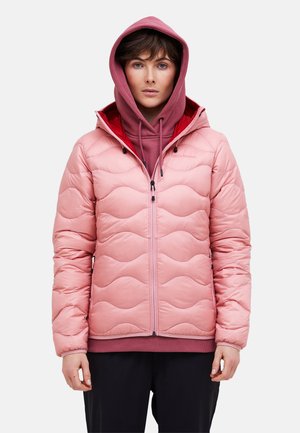 Jongere draagt een lichtroze gewatteerde jas over een poederroze hoodie met de capuchon op, staand tegen een effen achtergrond.