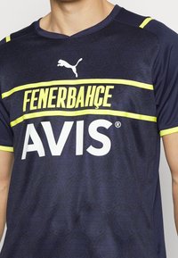 Laivastonsininen urheilupaita keltaisin yksityiskohdin; valkoisella lihavoidulla tekstillä "FENERBAHÇE" ja "AVIS"; kohokuvioitu kangas logokuvioilla.