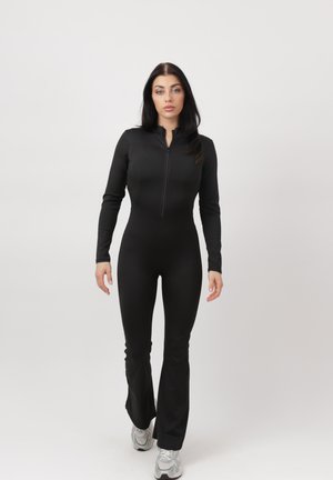 Kobieta o długich ciemnych włosach, ubrana w dopasowany czarny kombinezon z długim rękawem i białe sneakersy, stoi na tle jednolitego jasnego tła.