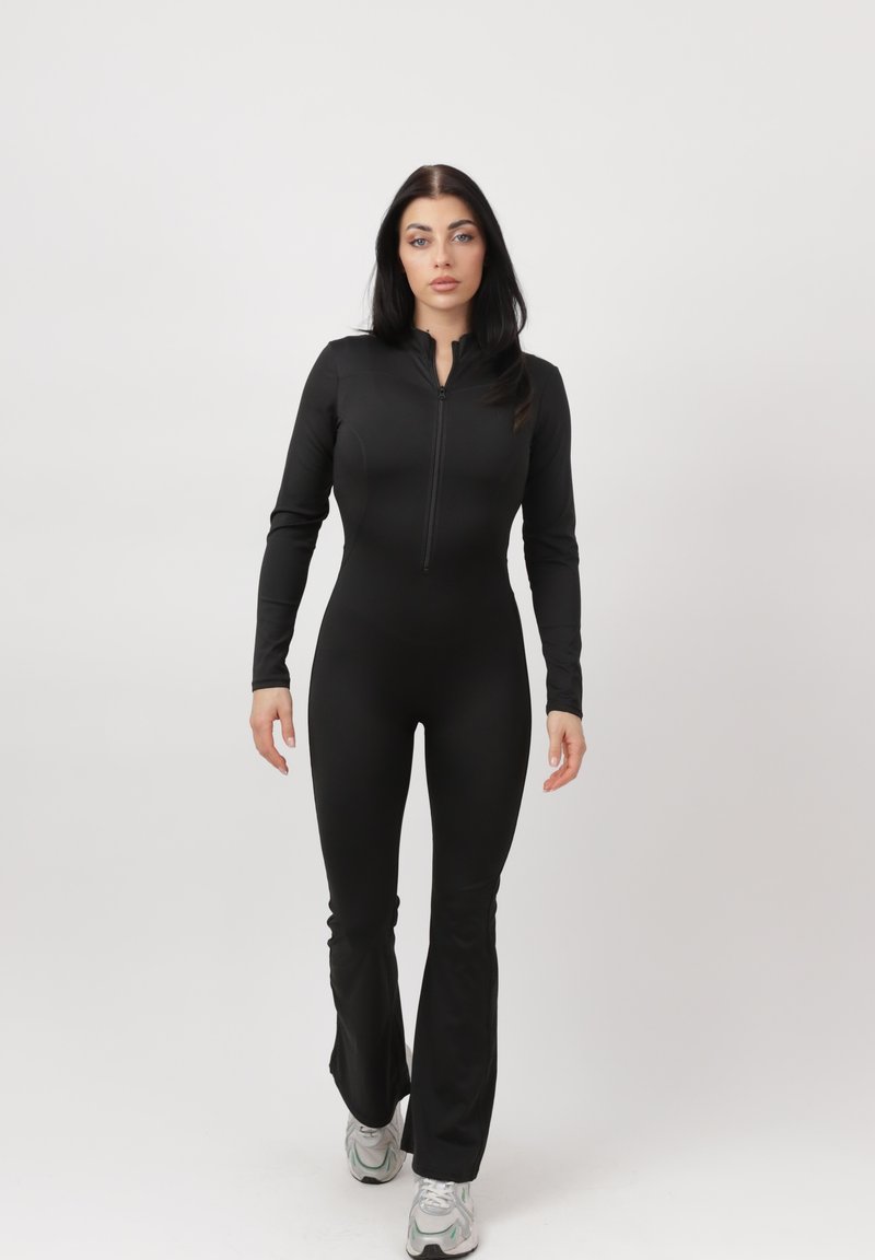 Mujer de cabello largo y oscuro viste un mono negro ajustado de manga larga y zapatillas blancas, de pie contra un fondo claro y liso.
