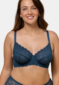Soutien-gorge en dentelle bleu profond avec des motifs floraux, tissu semi-transparent, bords ondulés et bretelles ajustables avec un bouton décoratif central.