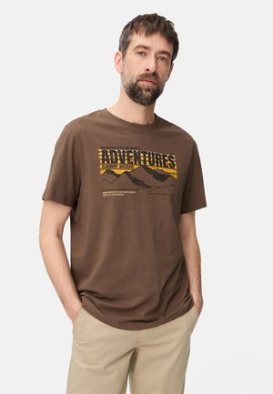 Mężczyzna z krótkimi włosami i brodą, ubrany w brązowy t-shirt z napisem "Adventures" oraz beżowe spodnie, stojący z rękami w kieszeniach na białym tle.