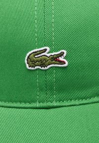 Gorra verde hecha de tela texturizada que presenta un logotipo de cocodrilo verde bordado con detalles rojos en el lado izquierdo.