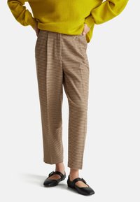 Bruine en beige houndstooth-gepatroond broek met een losse pasvorm, plooien en een verkorte lengte, gedragen met zwarte platte schoenen. Heldere gele trui.