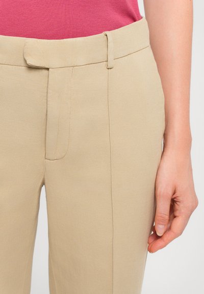 Pantalones beige de corte ajustado con frente plano, Passant para cinturón y bolsillos laterales, hechos de una tela lisa y texturizada. Combinados con una blusa rosa.